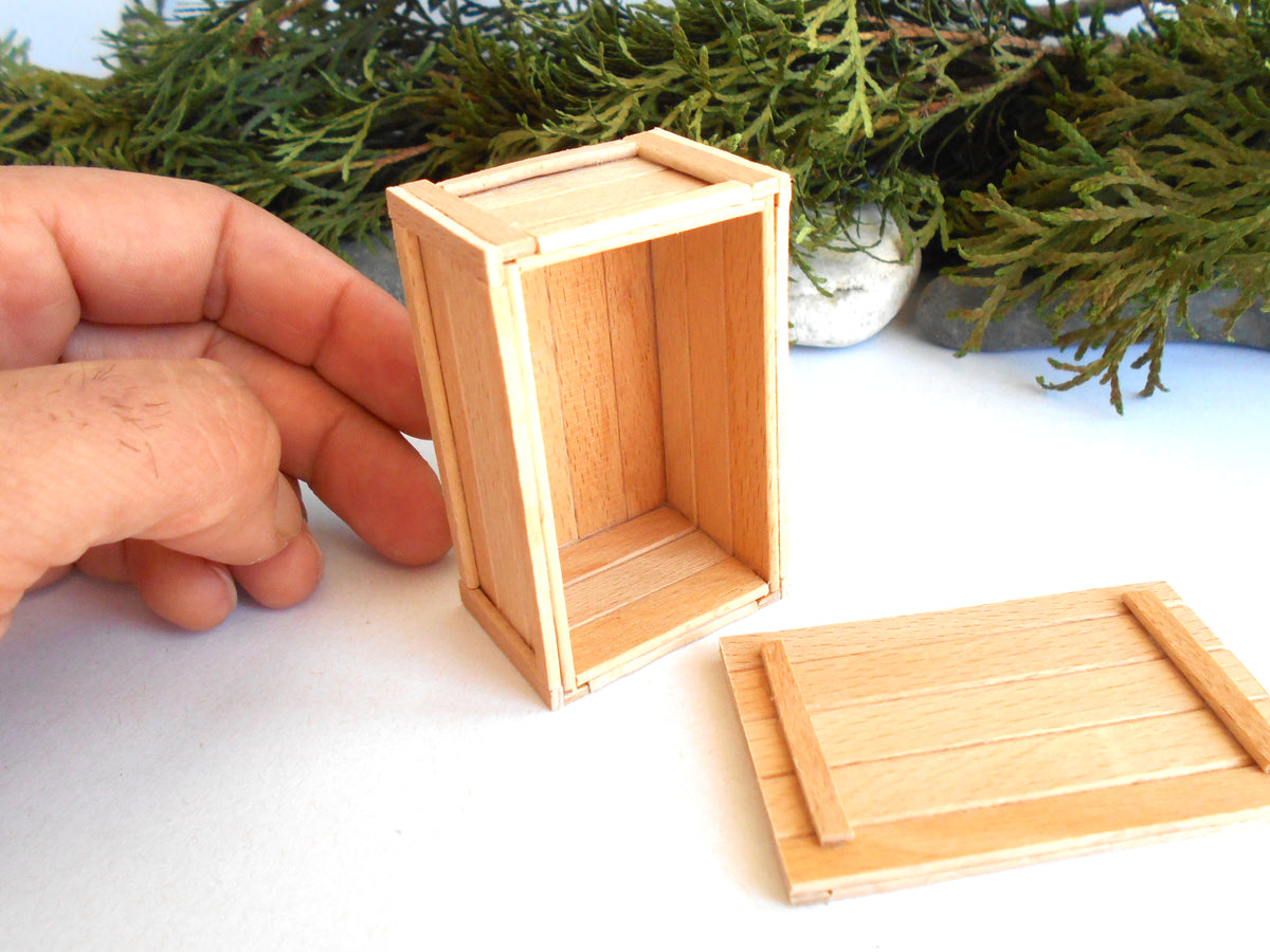 A miniature unfinished wooden crate box with its lid placed beside it, held by a hand to show scale, with green foliage in the background.
