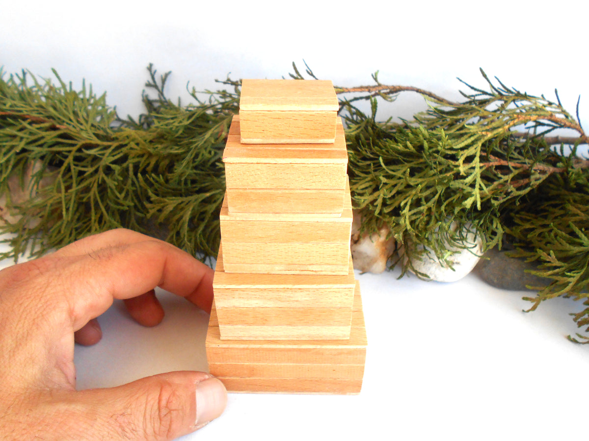 A hand holds a small wooden miniature boxes set with a natural light wood finish, positioned in front of a green evergreen garland on a white background.