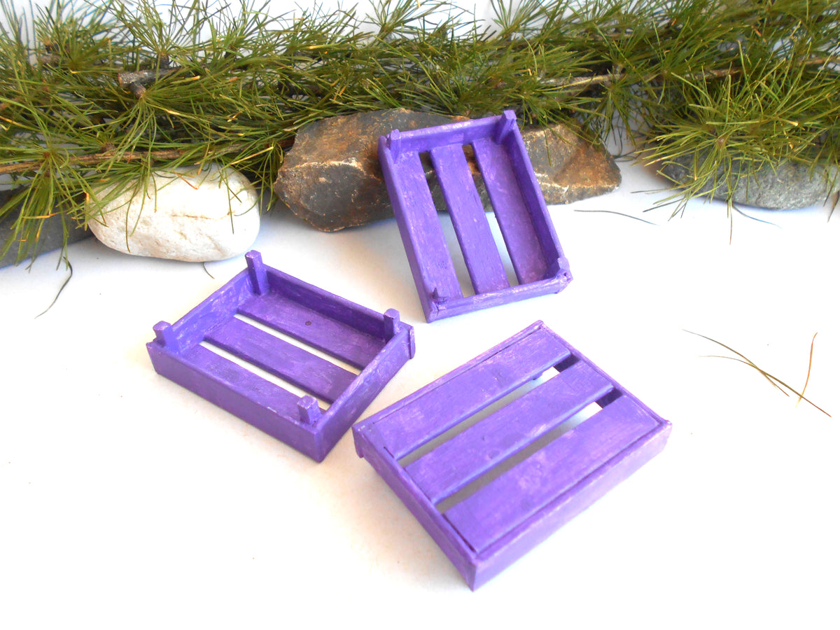 Three miniature purple wooden crate ornaments arranged on a white surface with decorative pine branches and small stones in the background.