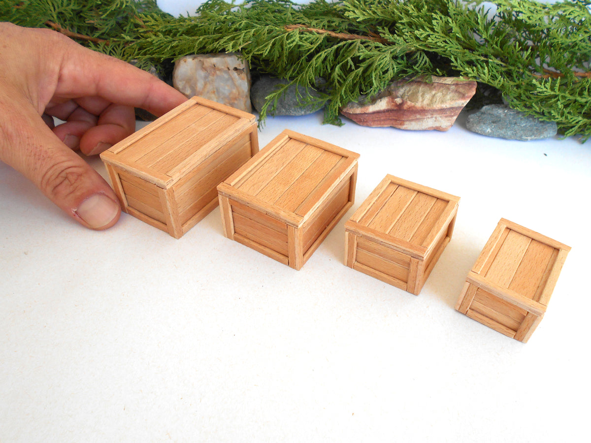 Four miniature light brown wooden crates of varying sizes arranged diagonally on a white surface, with a hand holding the largest crate and green foliage in the background.