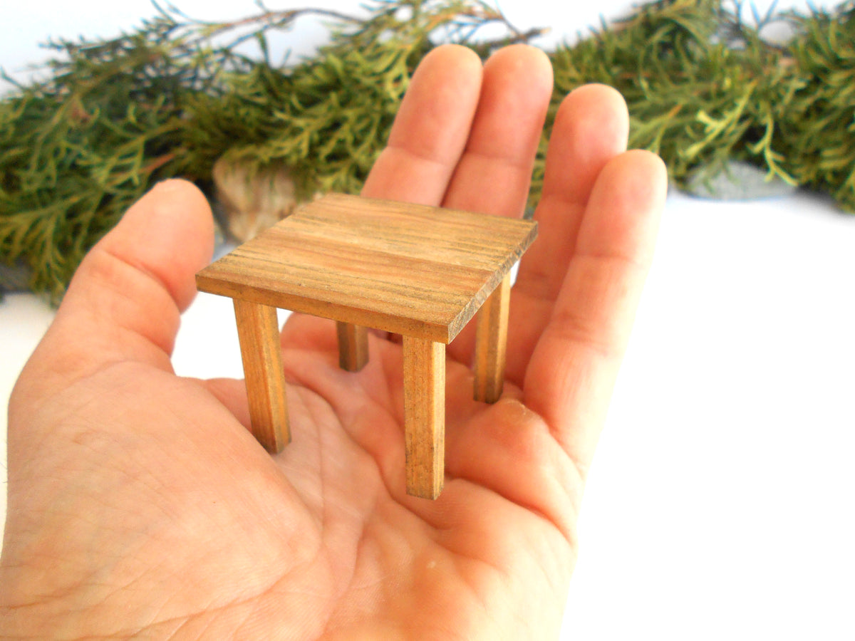 A miniature wooden dollhouse table held in a person's hand, with a blurred background of green foliage and white surface.