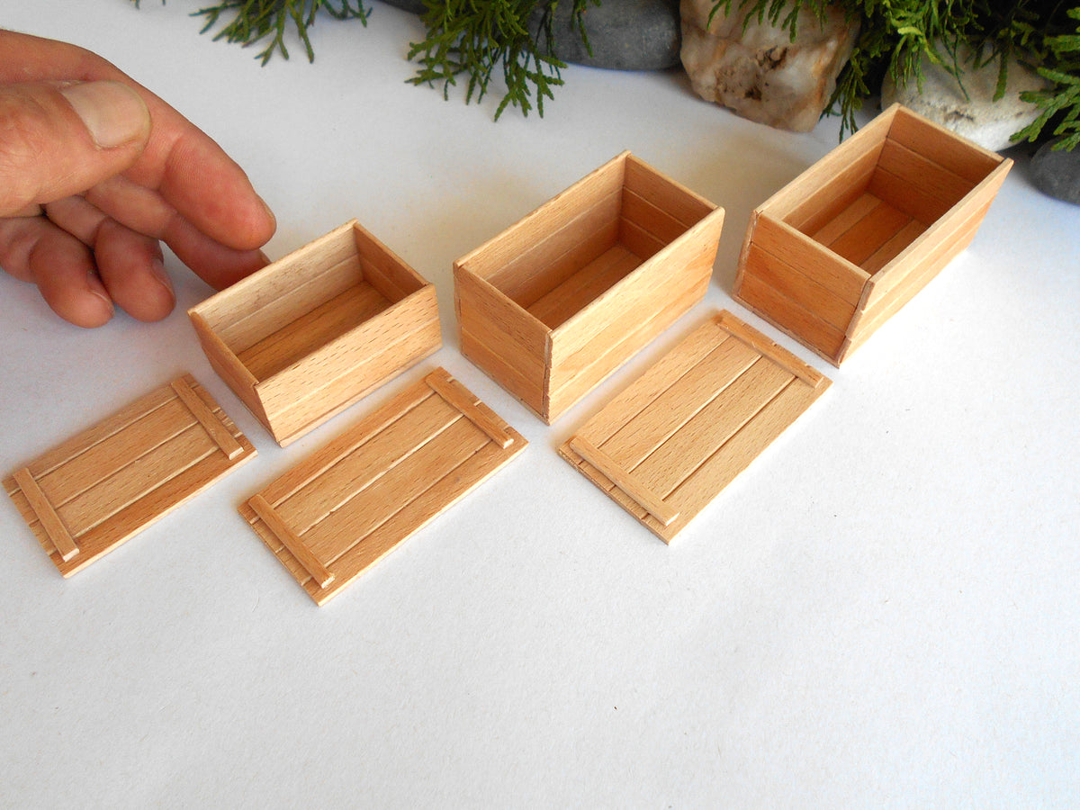 Six miniature light wood boxes and matching lids arranged on a white surface, with a hand pointing toward one of the small boxes. The background includes some blurred greenery and stones.