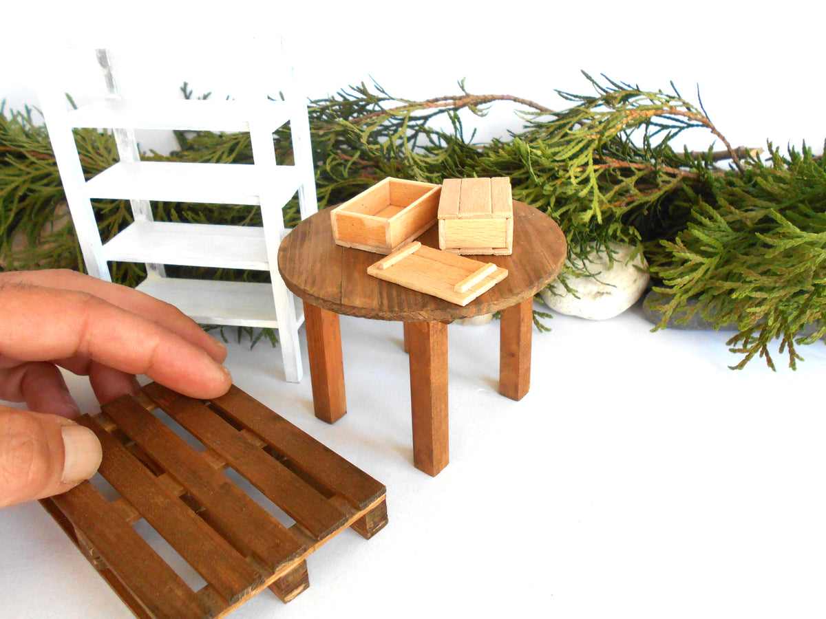 A miniature wooden pallet table component being held by a hand, with a miniature round wooden table, small wooden boxes, and a white miniature shelving unit in the background.