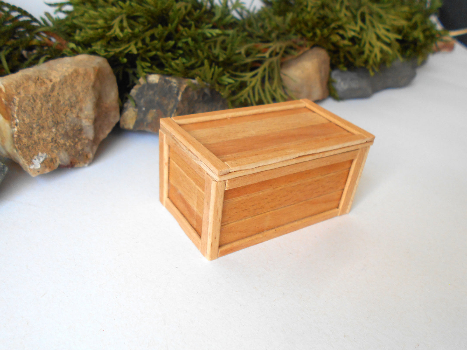 A miniature light wood box with a hinged lid, shown on a white surface with a person's fingers visible at the bottom for scale, and green foliage and small rocks in the background.