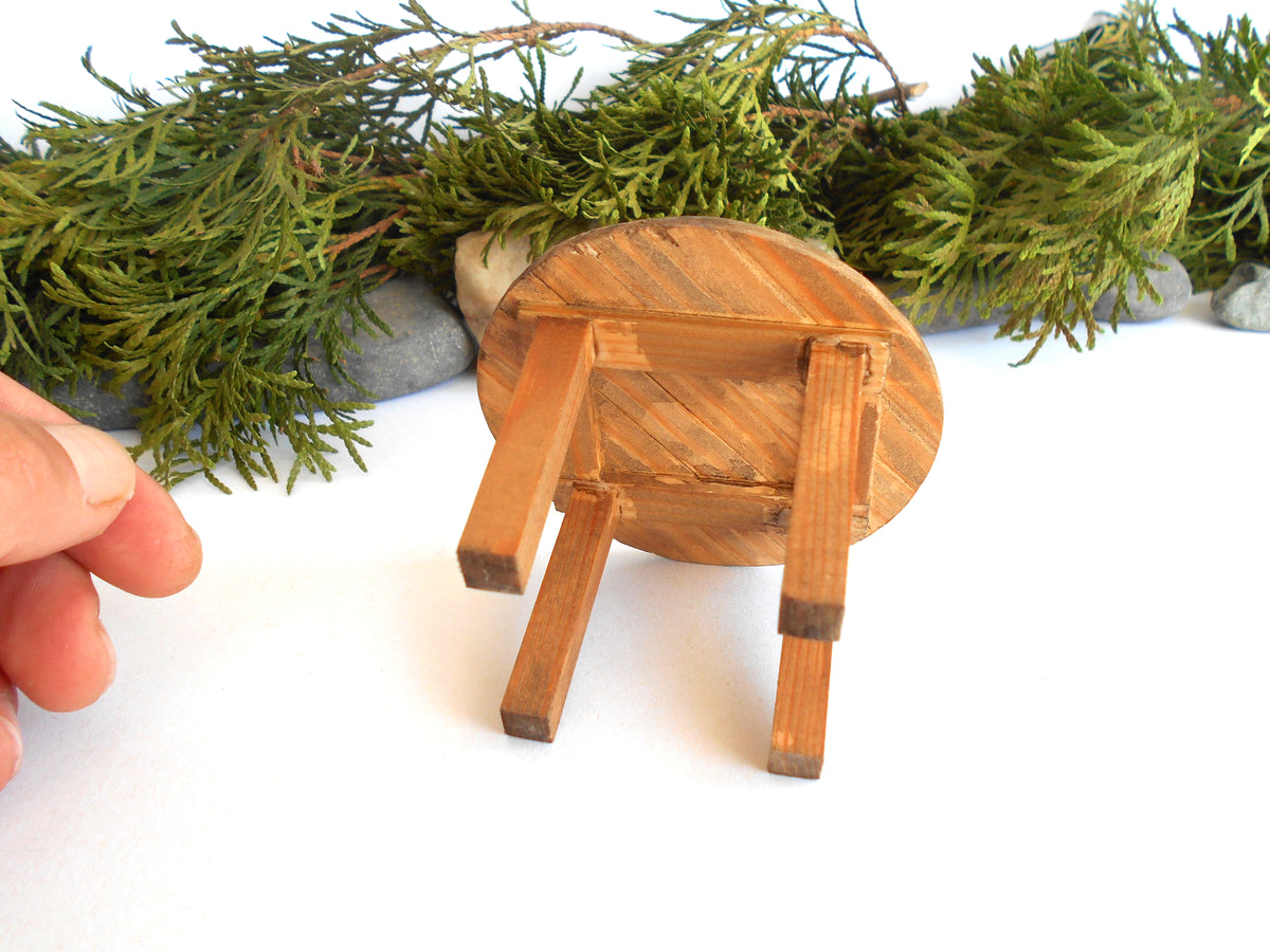 A miniature round wooden stool or small stool prop shown on a white surface with green foliage and stones in the background, and a human hand entering from the left to indicate scale.
