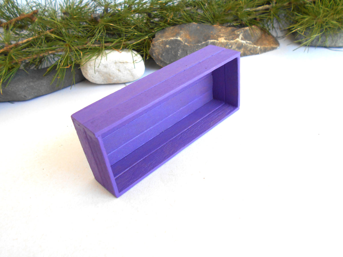 A small purple wooden shelf or display box with an open top, photographed on a white surface with pine branches and rocks in the background.