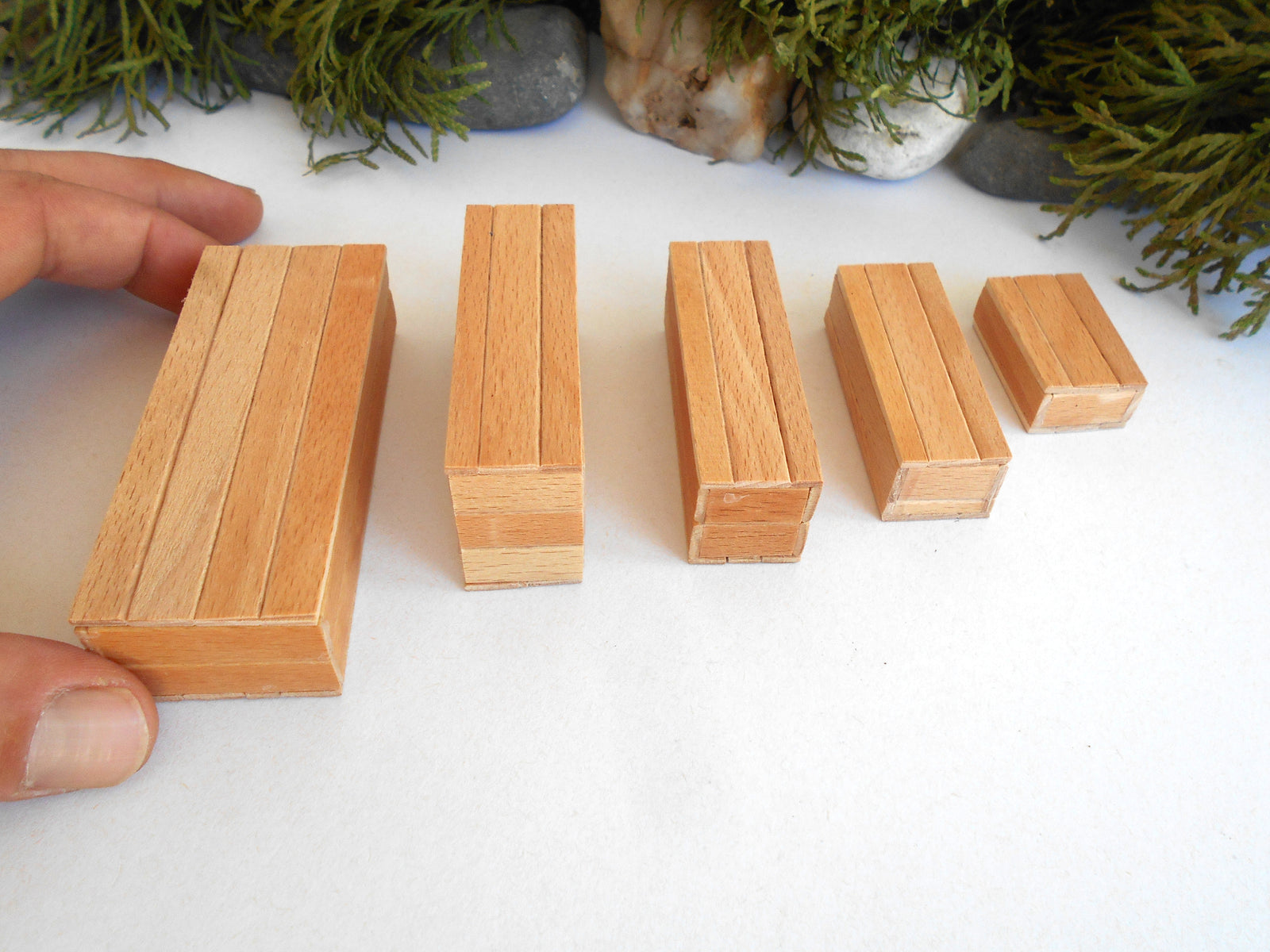 Five rectangular wooden miniature boxes of varying sizes arranged in descending size order on a white surface, with a hand holding one of the larger blocks on the left. Green foliage and stones are visible in the background.