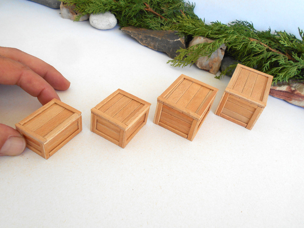 Four miniature light brown wooden crates arranged in a row on a white surface, with a person's hand touching the leftmost crate and greenery in the background.