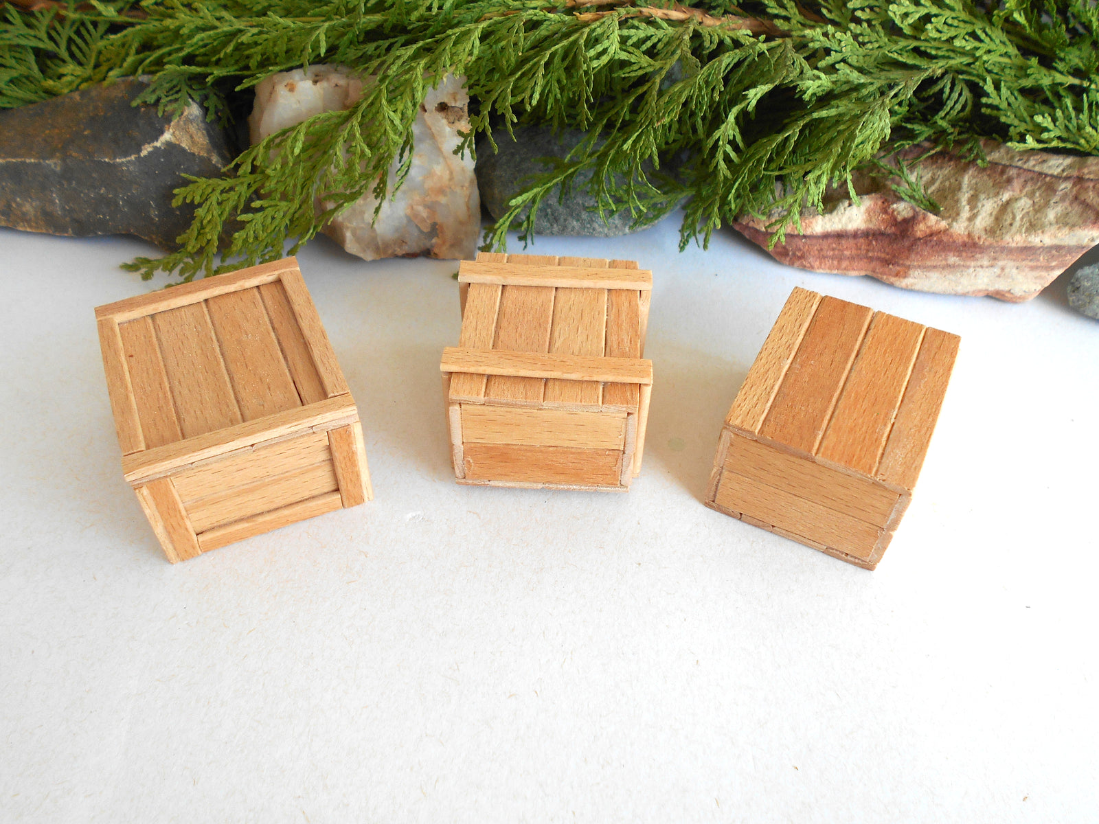 Three miniature wooden crates in different positions, displayed on a white surface with green foliage and rocks in the background.