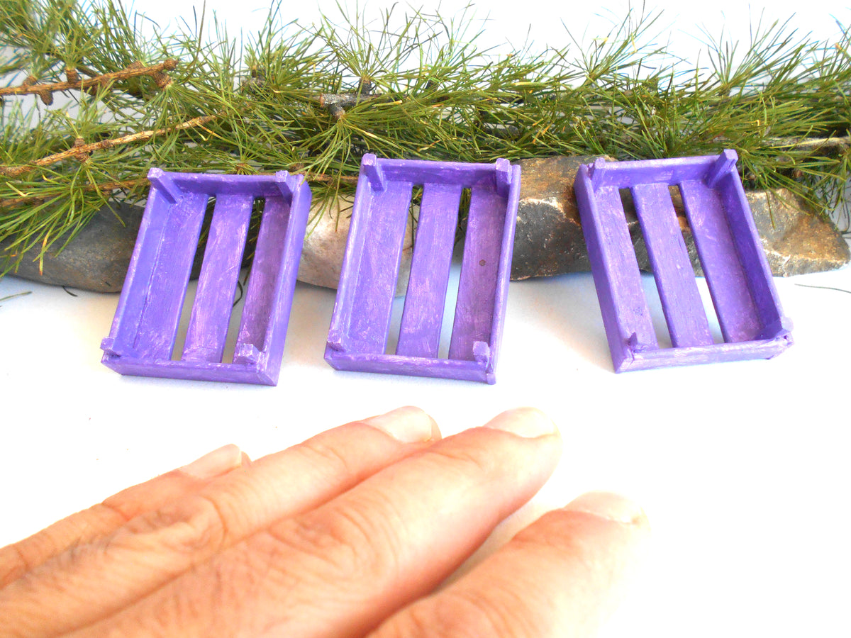 Three miniature purple plastic crate accessories arranged on a white surface with pine branches in the background and a person's hand visible in the foreground for scale.