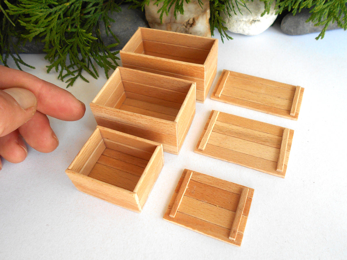 Six miniature light wood boxes and trays, including three open square boxes and three rectangular lids, displayed on a white surface with a hand pointing to one of the boxes for scale.