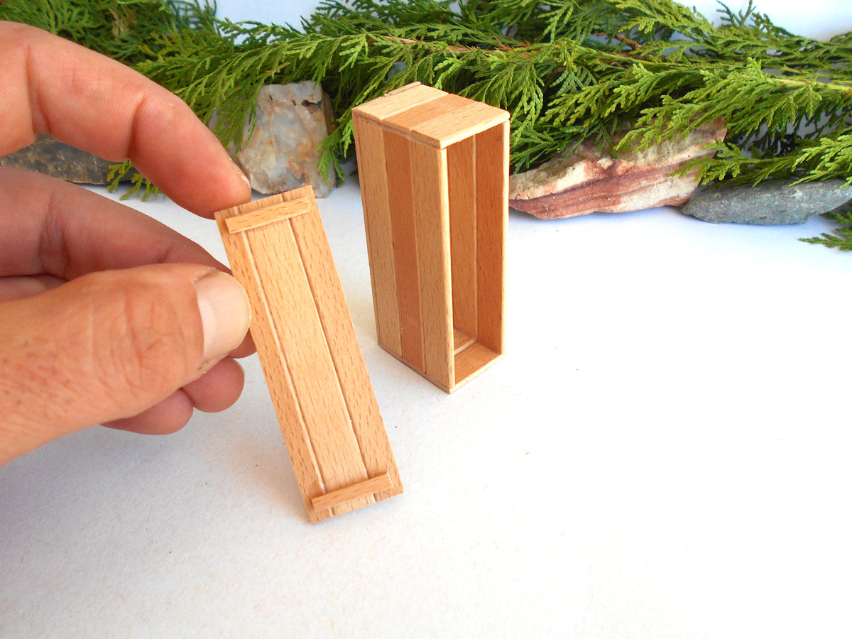 A hand holds a small light wood box cover next to a matching open wooden box on a white surface, with green foliage and stones in the background.