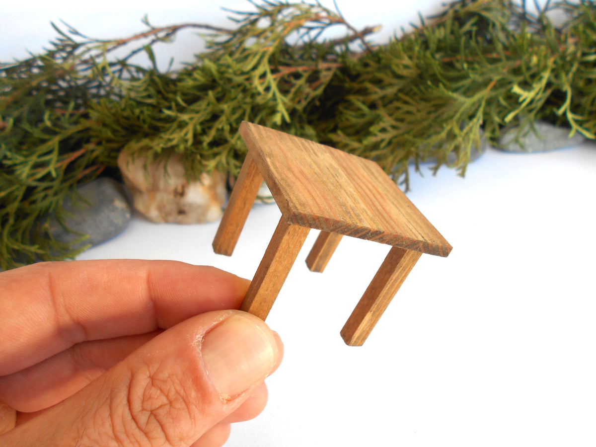 A miniature wooden end or side table held between fingers to show scale, with green foliage and stones blurred in the background on a white surface.