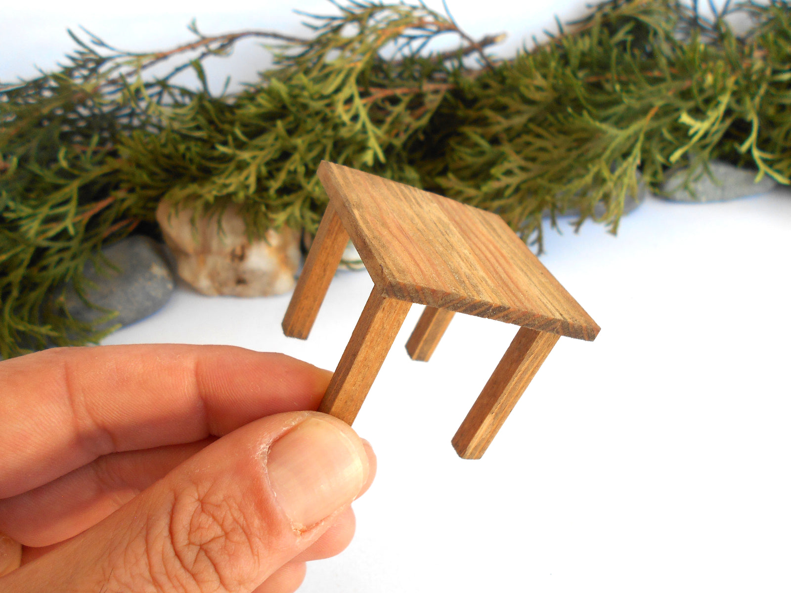 A miniature wooden end or side table held between fingers to show scale, with green foliage and stones blurred in the background on a white surface.