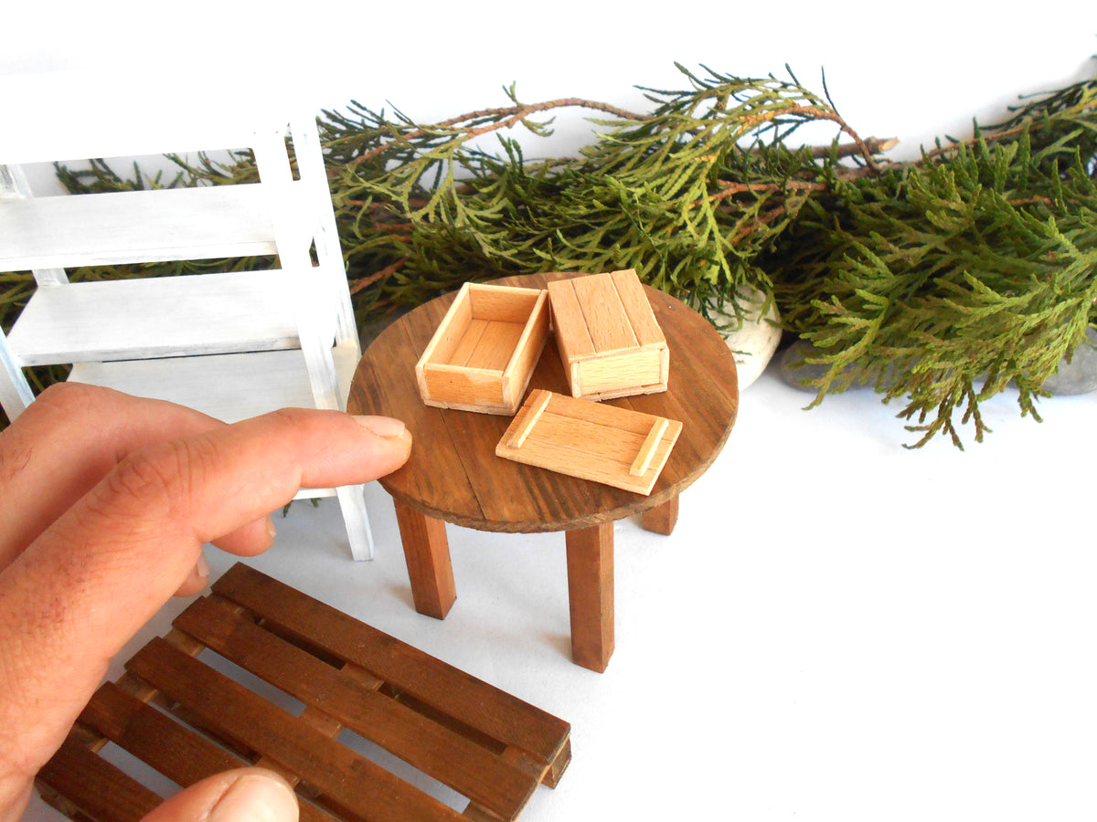 A miniature wooden dollhouse furniture set featuring a small round table with three open storage boxes, shown with a hand pointing at it. The scene includes a white chair, green foliage, and a wooden bench on a white background.