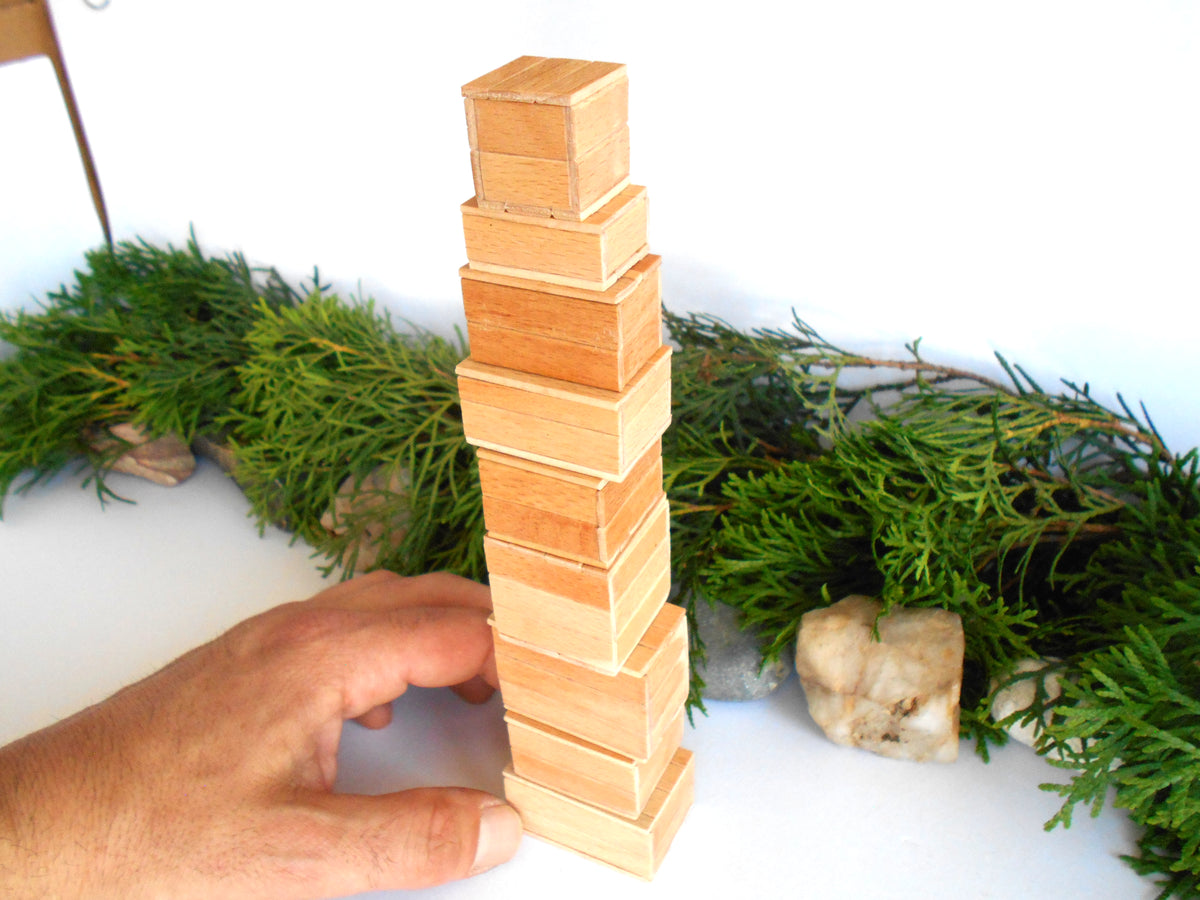 A hand is stacking natural wood boxes in a tower, with green garland and small stones arranged on a white surface against a white background.