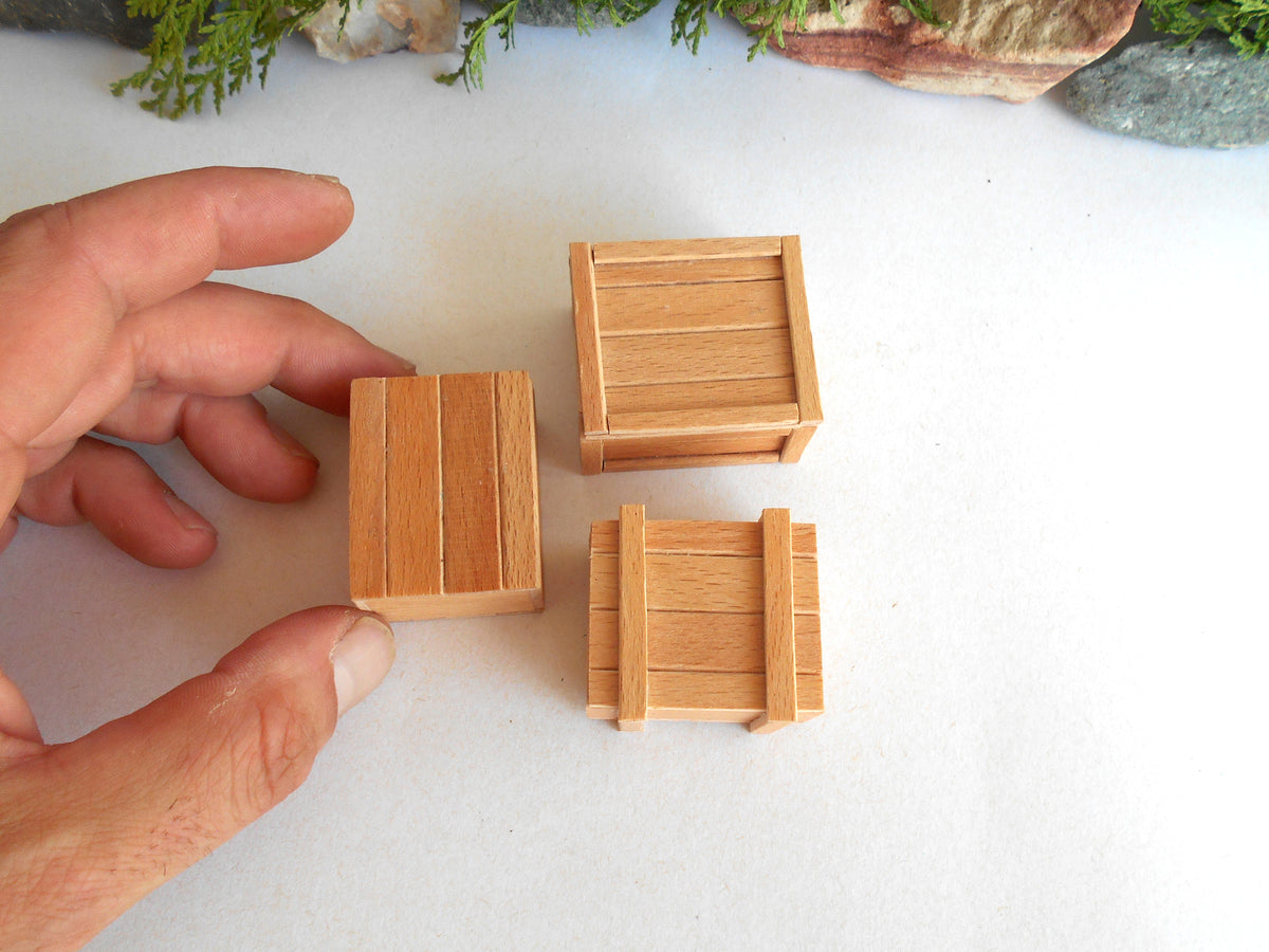 Three miniature wooden crates in different sizes, with one held by a hand to show scale, on a white surface with small decorative rocks and greenery in the background.