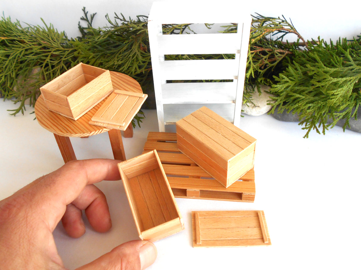 A hand holds a miniature wooden crate in the foreground, with several matching small wooden storage boxes and crates arranged on a white surface. A small round wooden table, a white slatted chair, and green foliage are visible in the background.