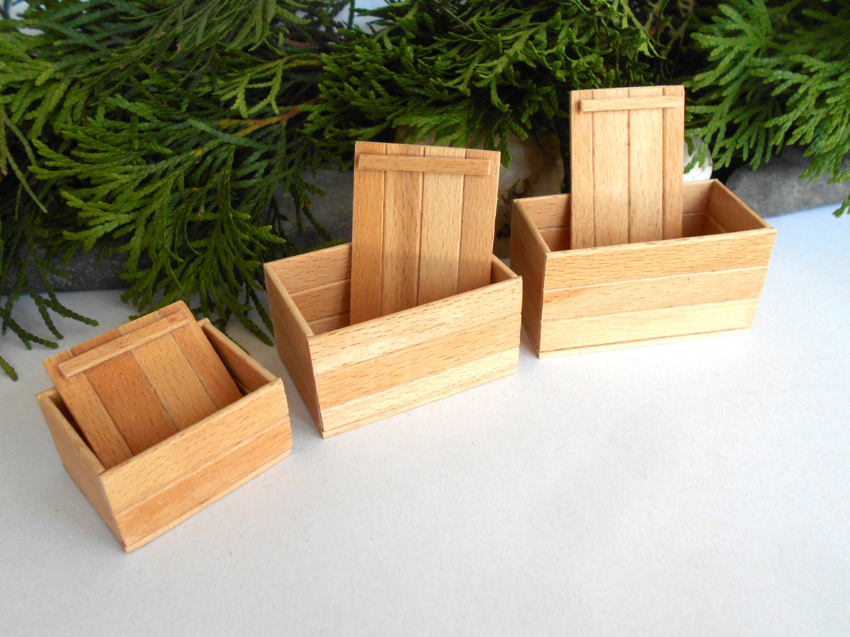 Three small light wood boxes with slatted lids, arranged in ascending size on a white surface with green foliage in the background.