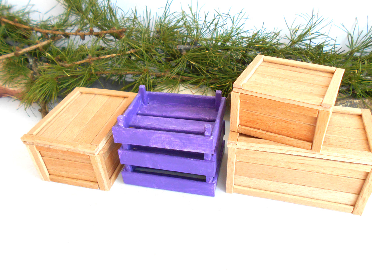 Three miniature wooden crates of different sizes, including two natural light wood crates and one painted purple crate, arranged on a white surface with green pine branches in the background.
