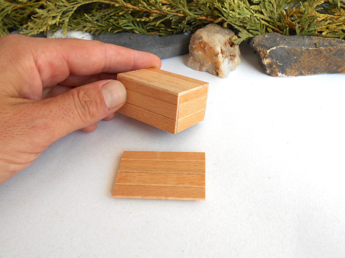 A hand holds a small rectangular wooden box lid next to its matching base on a white surface, with decorative greenery and stones in the background.