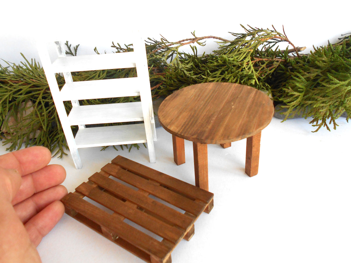 A miniature dollhouse furniture set featuring a round wooden table, a wooden pallet-style tray, and a white shelving unit, with a hand holding the tray in the foreground against a white background with green garland.