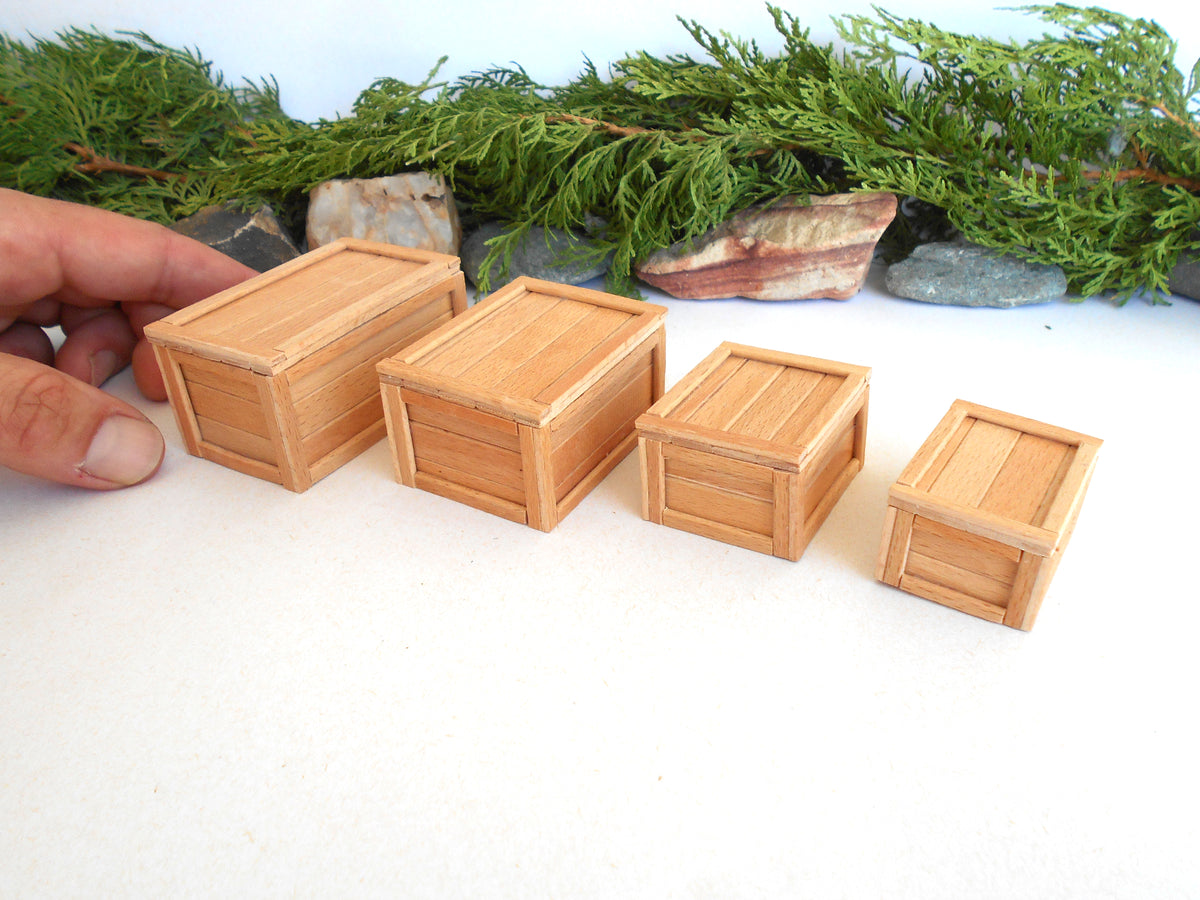 Four miniature light wood crates of varying sizes arranged on a white surface, with a hand holding one of the larger crates and green foliage in the background.
