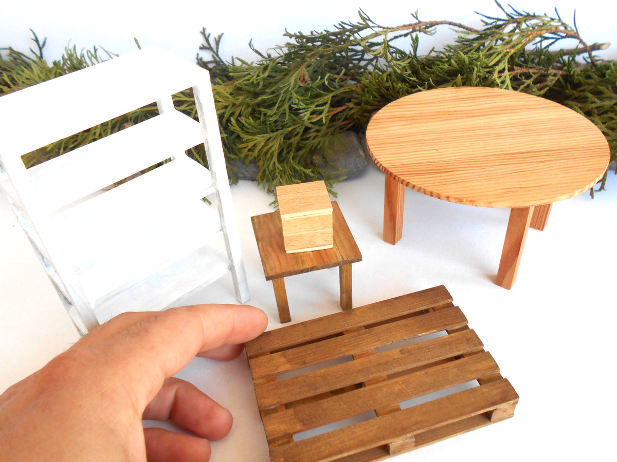 A miniature dollhouse furniture set featuring a round light wood table, a small square wooden stool with a matching box, a brown slatted pallet-style base, and a white shelving unit, with a hand holding the pallet in the foreground.