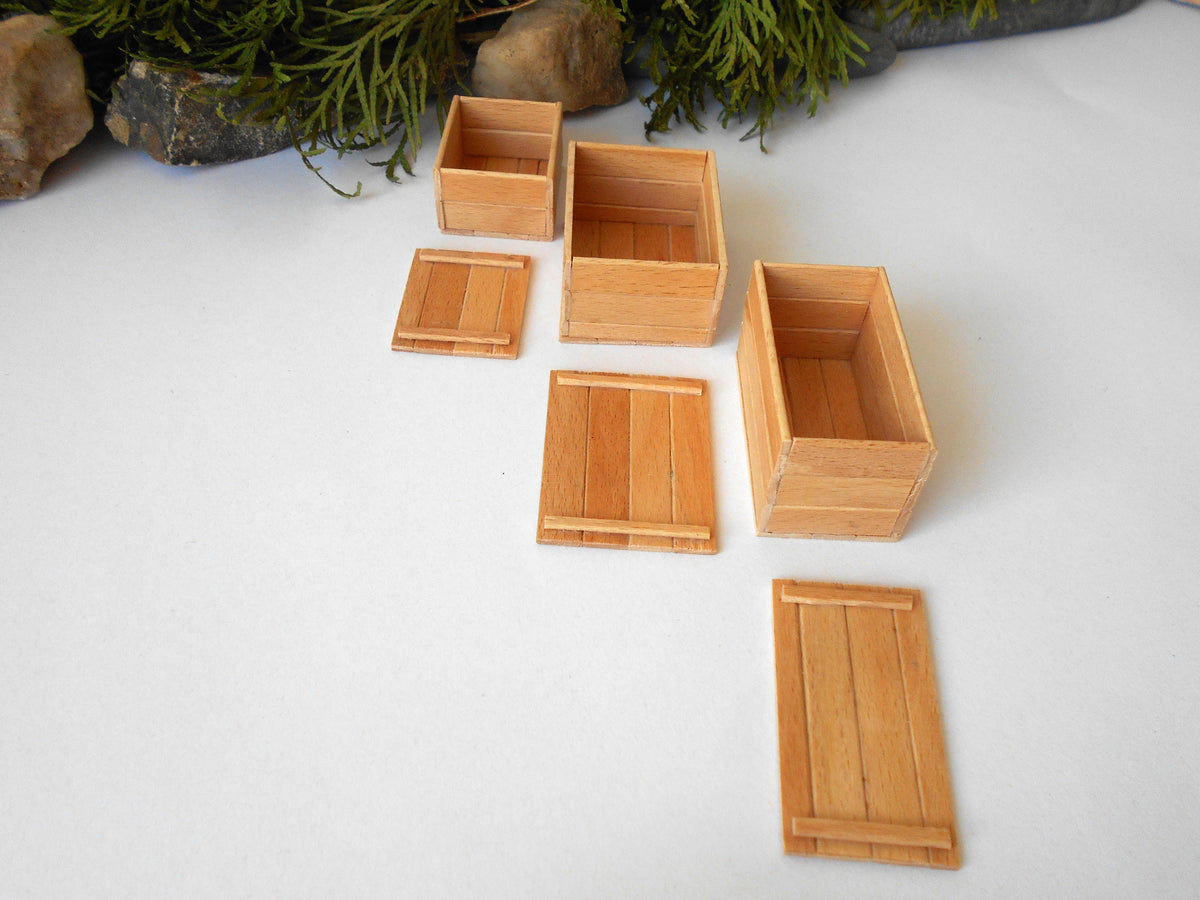Six miniature light brown wooden boxes and lids arranged on a white surface with decorative greenery and stones at the top.