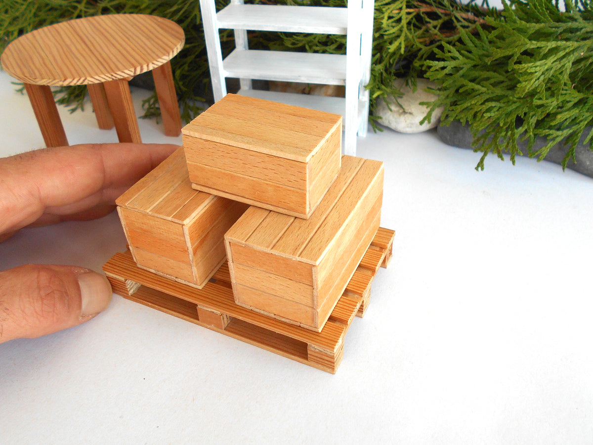 Three small light wood boxes stacked on a miniature wooden pallet, with a human hand holding the left side for scale, against a white tabletop with miniature furniture and greenery in the background.