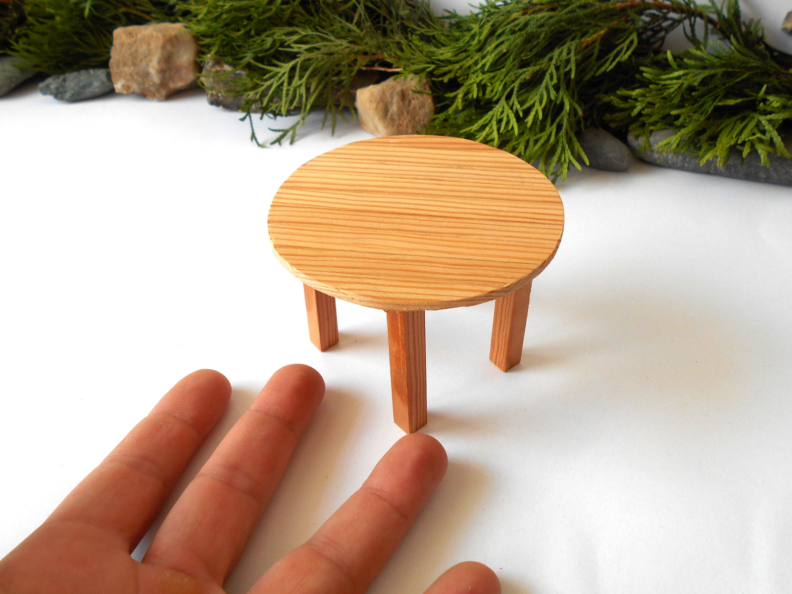 A miniature round wooden stool or table with three legs, shown next to a human hand for scale on a white surface with green foliage in the background.