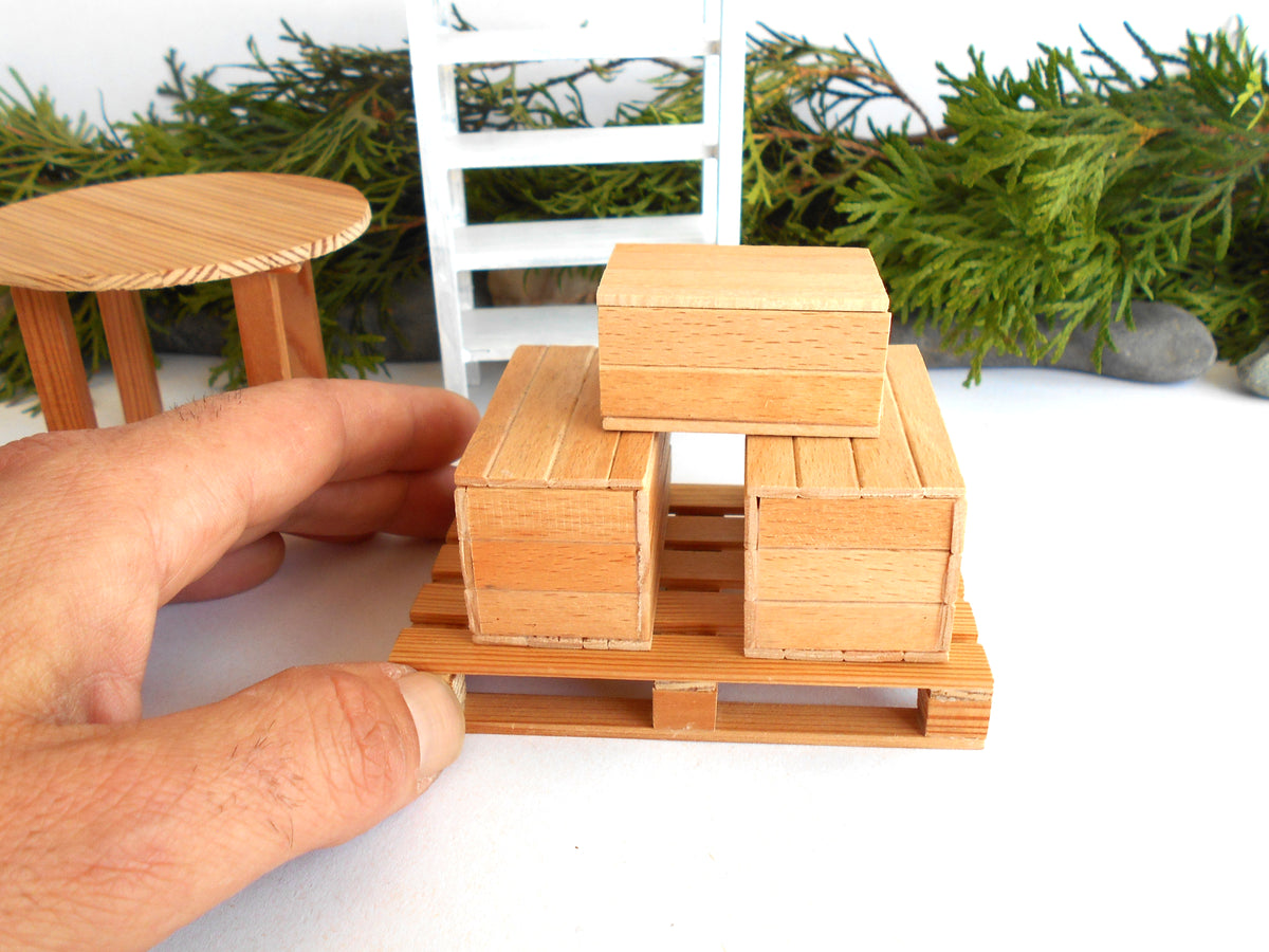A miniature wooden crate set on a small wooden pallet, with a hand holding it for scale, against a white background with miniature furniture and greenery.