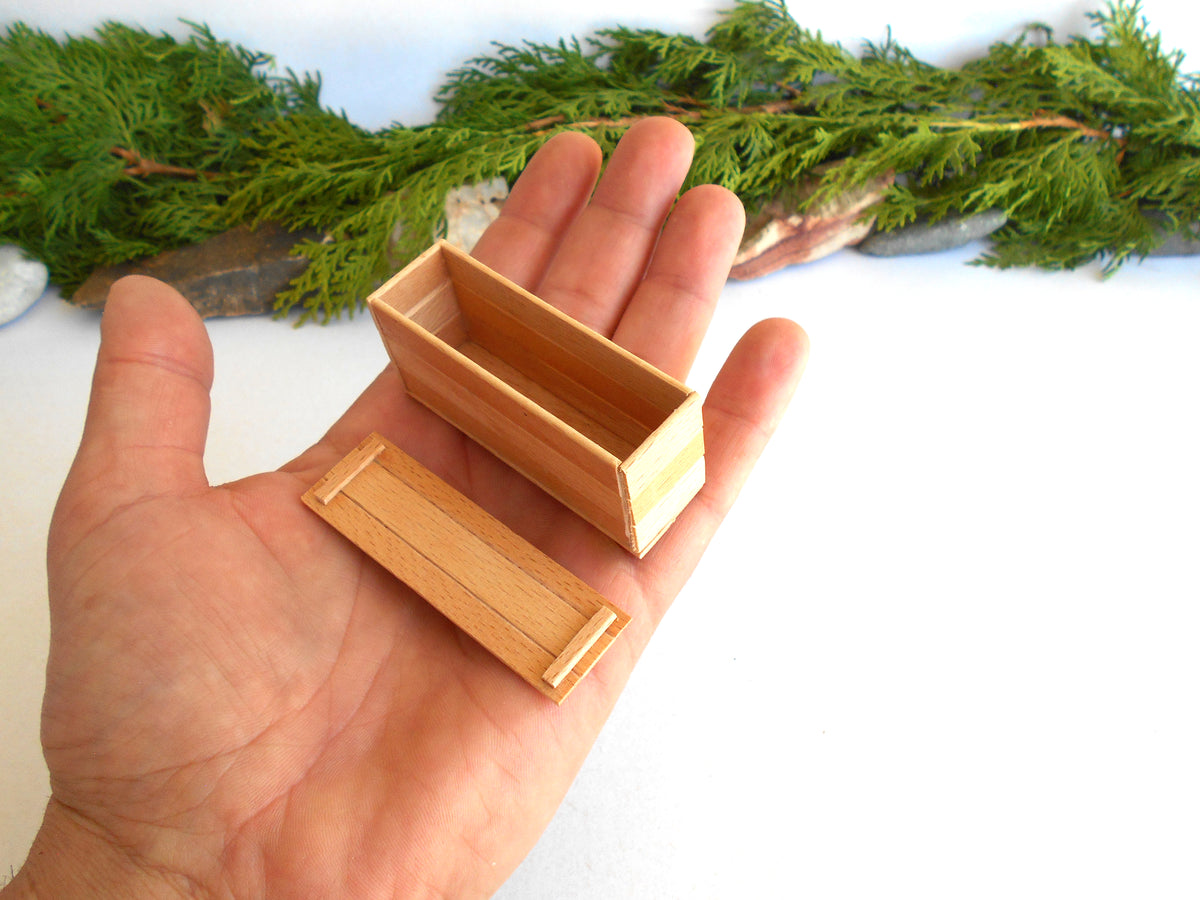 A miniature light wood box with its lid placed beside it, held in a person's hand. The background features a white surface with a decorative green pine garland.