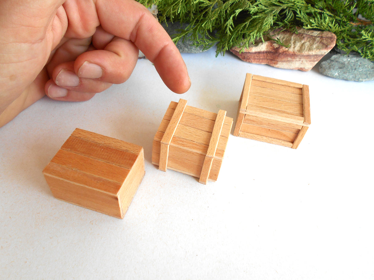 Three miniature light wood boxes of different designs on a white surface, with a person's hand pointing at the center box and green foliage in the background.