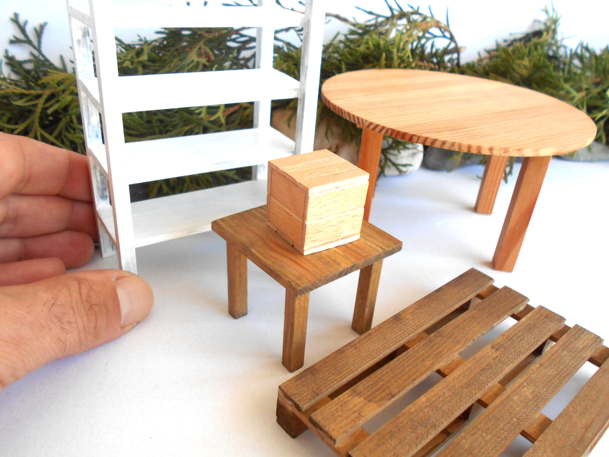 A miniature dollhouse furniture set including a round wooden table, a small wooden stool with a box on top, a wooden pallet, and a white shelving unit, shown with a hand for scale.