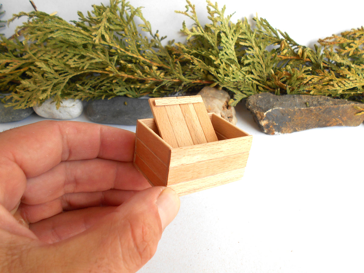 A miniature light wood box with a slatted lid, held in a person's hand against a white background with decorative greenery and stones.