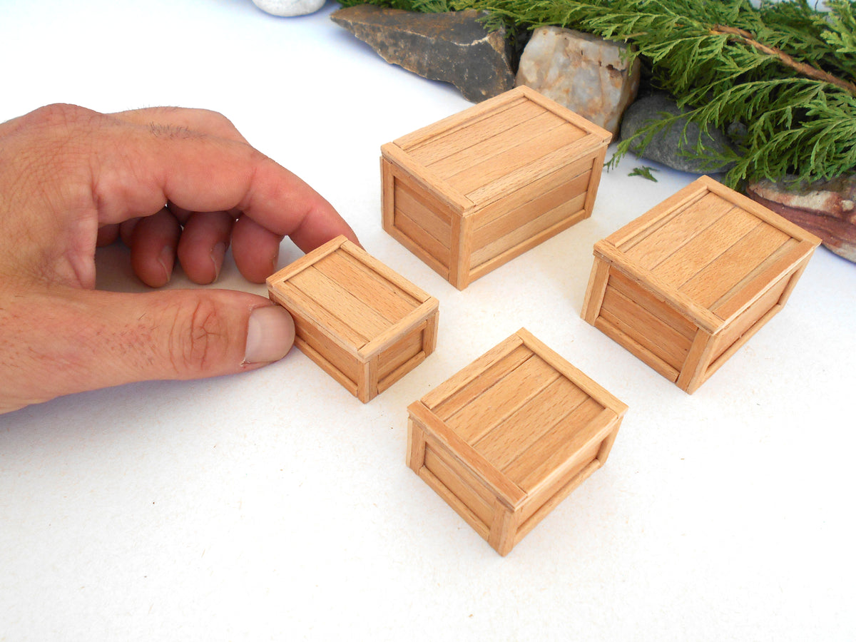Four miniature light brown wooden crates of different sizes arranged on a white surface, with a hand holding one of the smaller crates to show scale. Green foliage and rocks are visible in the background.