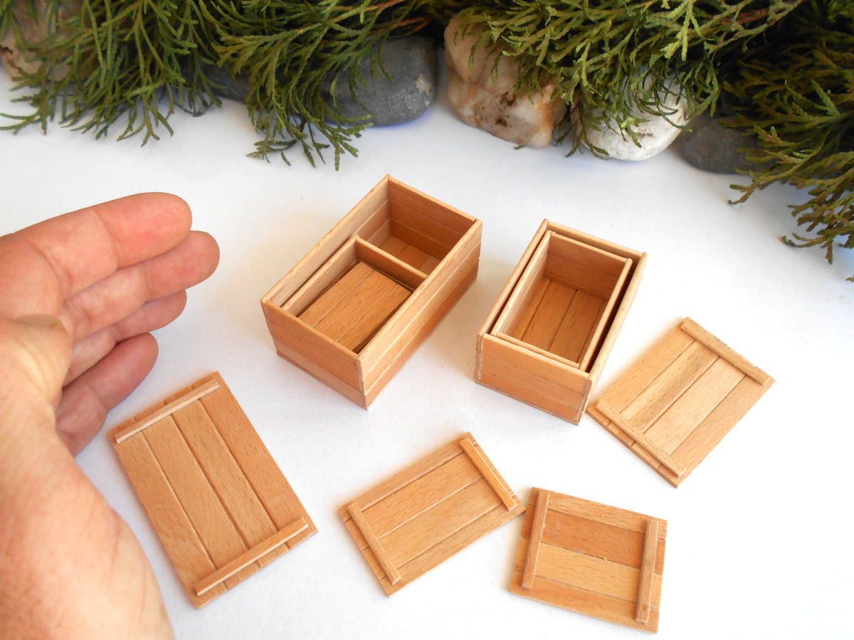 Six miniature light wood boxes and lids arranged on a white surface, with a hand visible on the left for scale and greenery in the background.