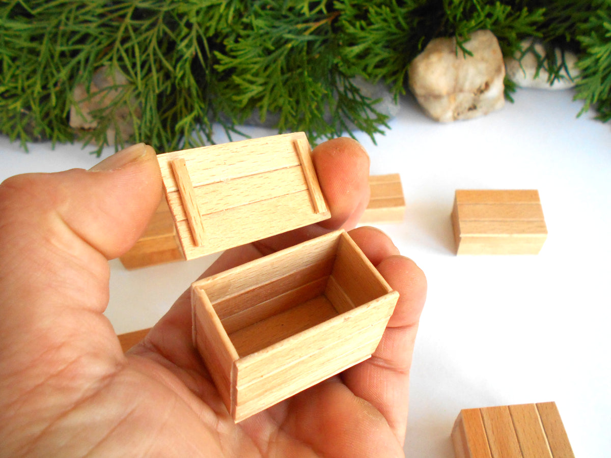 A hand holding a small wooden box with its lid partially open, with other miniature wooden boxes and green foliage in the background.