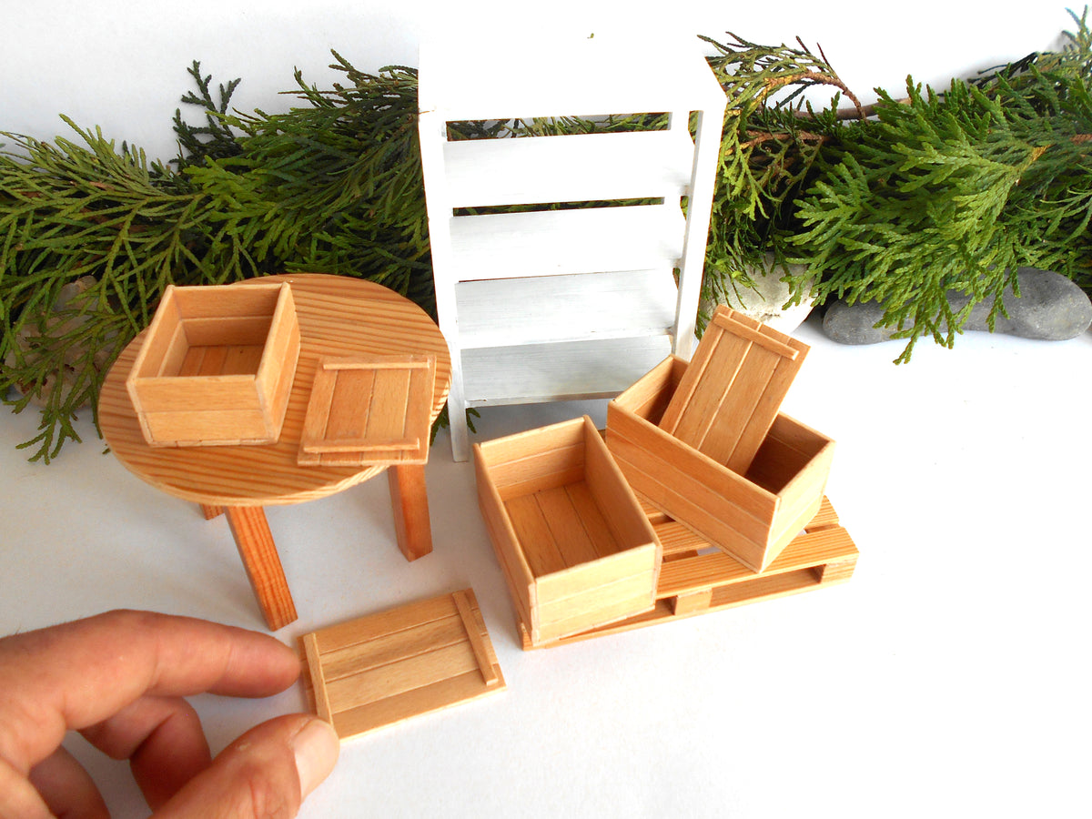 A set of miniature wooden dollhouse furniture pieces, including a round table, shelving unit, and various storage crates, shown with a hand holding a small crate to indicate scale against a white background with green pine branches.
