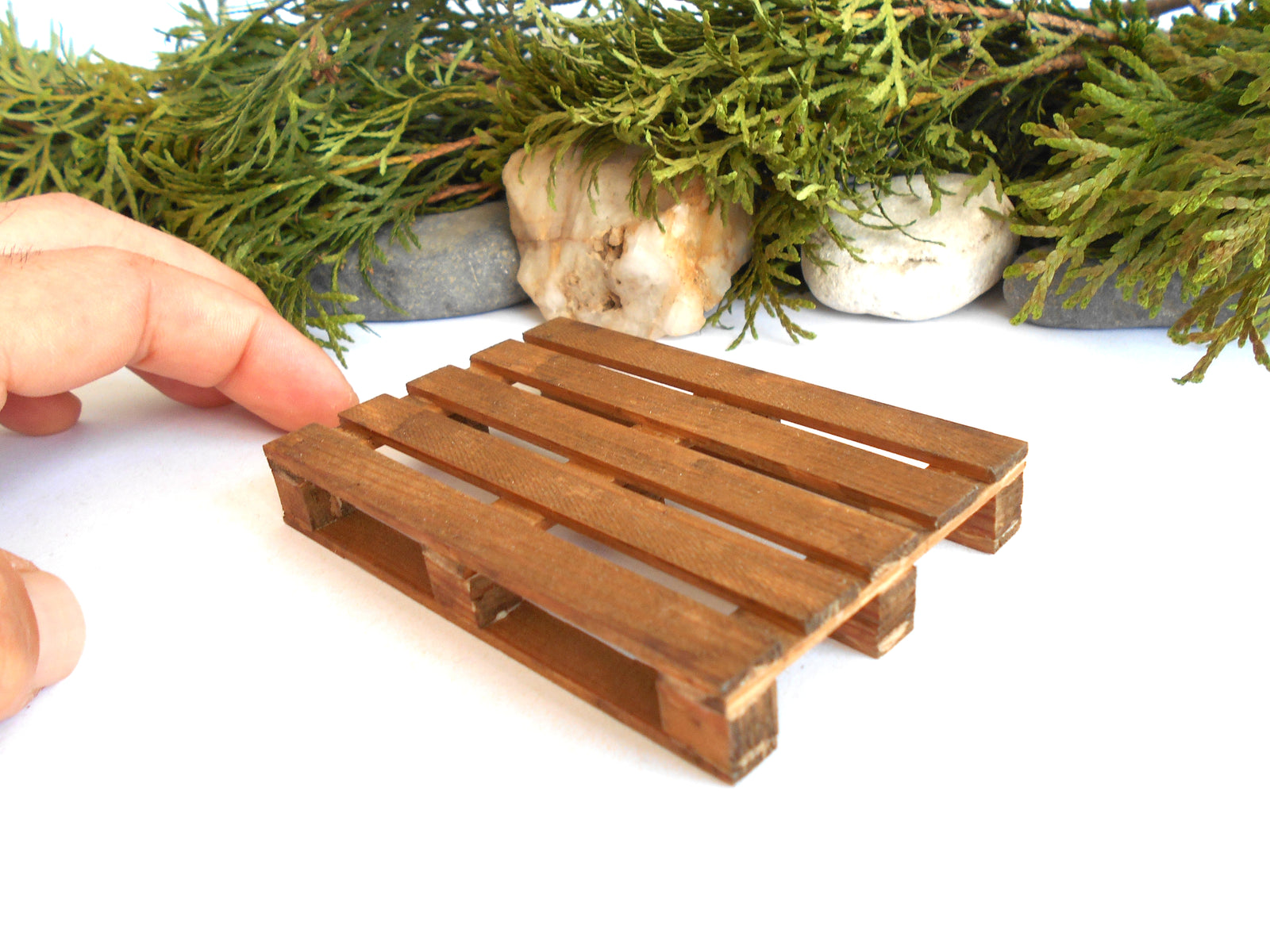 A miniature wooden pallet model being held by a hand, set against a white background with decorative green foliage and stones.
