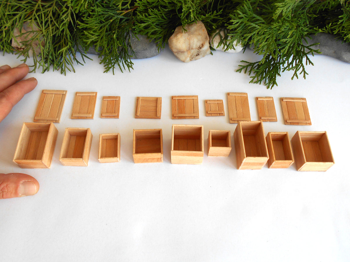 A set of miniature light wood boxes and crates in various sizes, arranged on a white surface with green foliage at the top and fingers visible on the left side.