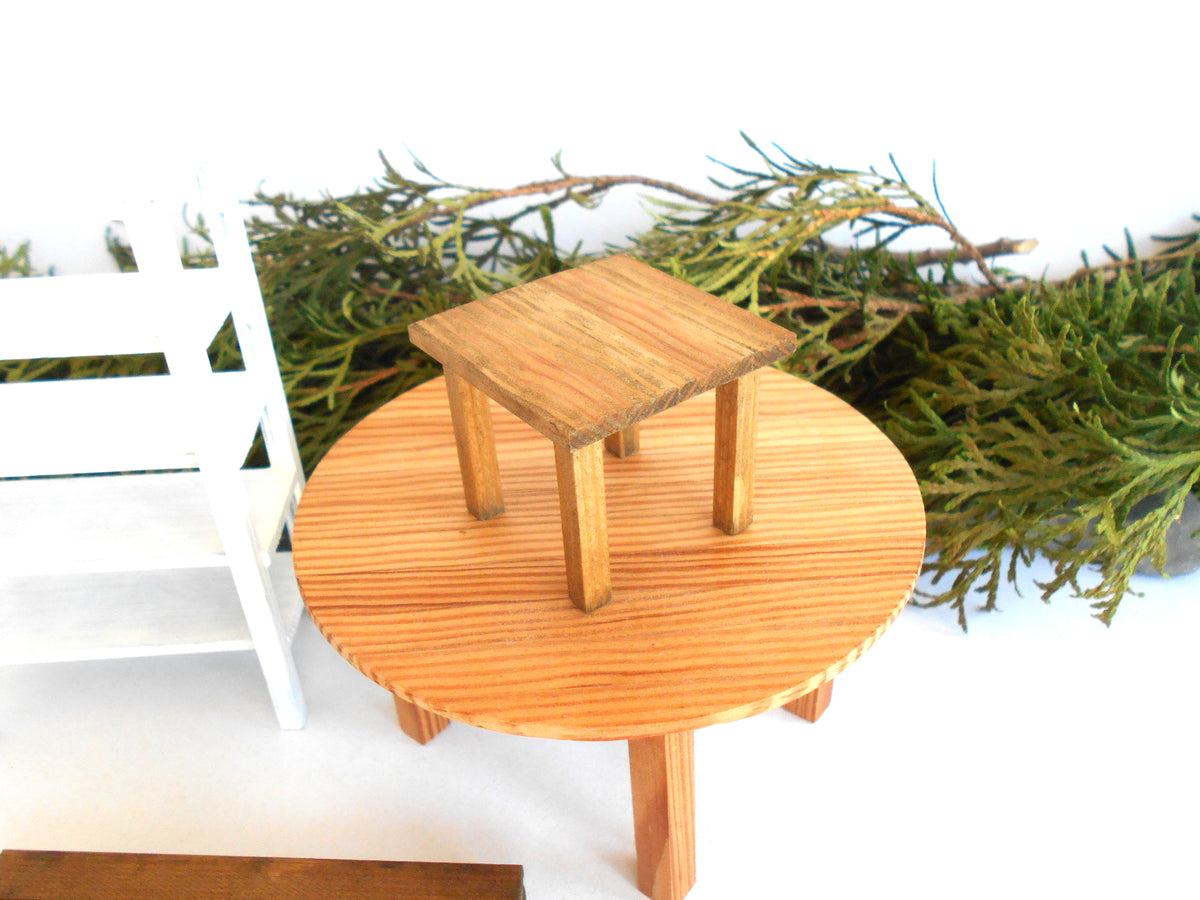 A miniature wooden side table with a round light brown tabletop and a small square wooden stool on top, set against a white background with green foliage and a white chair partially visible.