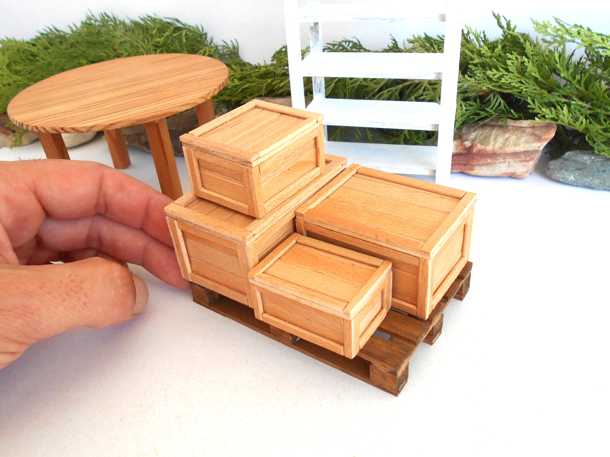 A hand holding a miniature wooden crate set on a small pallet, with a dollhouse-style round table and white shelving in the background.