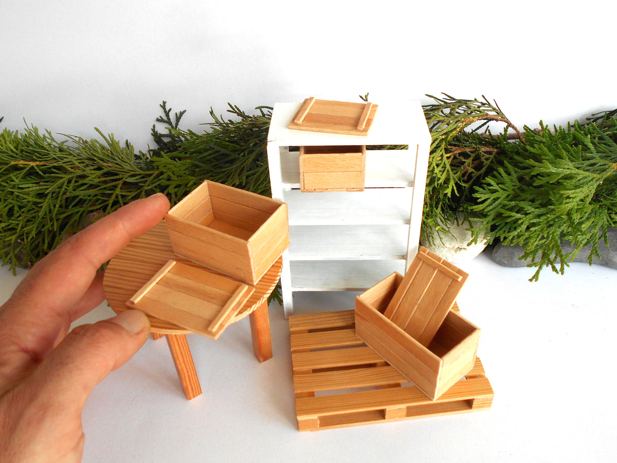 A miniature dollhouse furniture set made of light natural wood, including a small table, chairs, and shelving unit, shown with a hand holding one piece against a white background with green foliage.