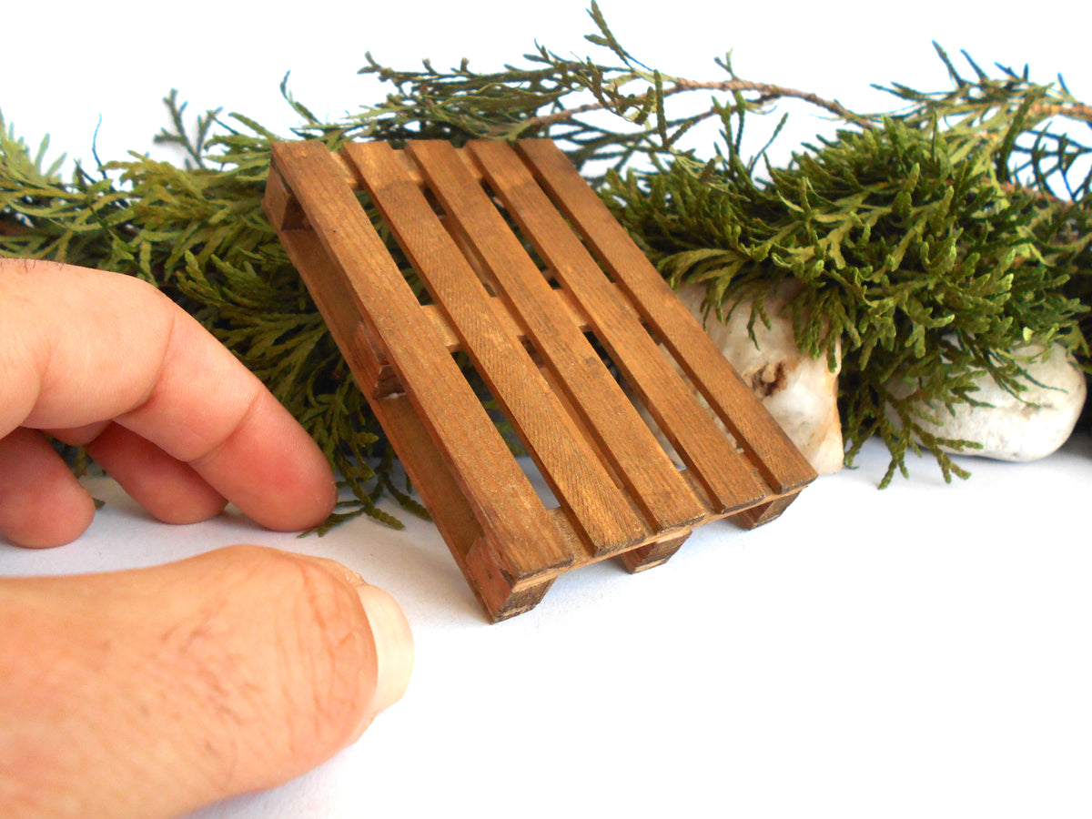 A miniature wooden slat tray or miniature pallet-style decor piece shown on a white background with greenery and a human hand holding it for scale.