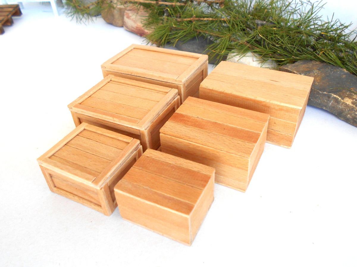 Six small rectangular wooden boxes with lids, arranged on a white surface with green pine branches and dark logs in the background.