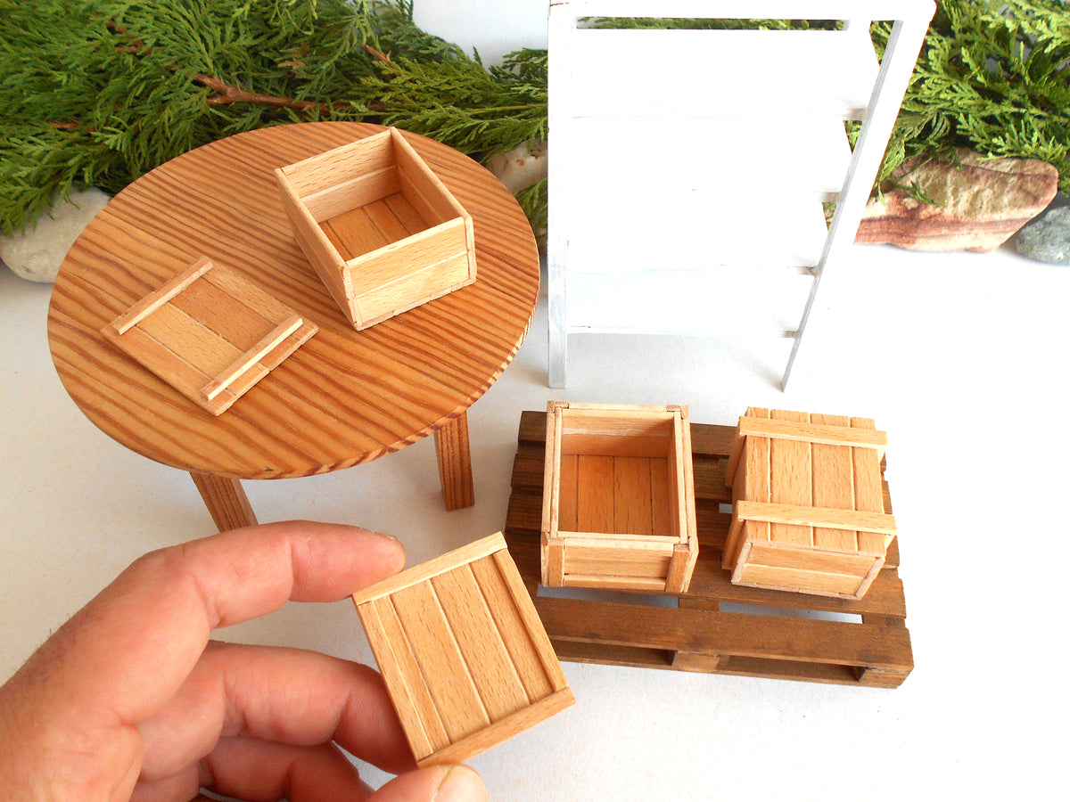 A miniature wooden crate set for dollhouse or model building, shown with a hand holding one crate in the foreground, a round wooden table, and a white shelving unit in the background.