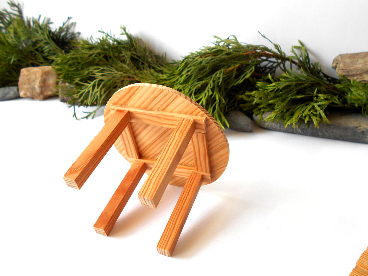 A small handcrafted wooden stool with a round seat and three legs, shown on a white surface with green foliage and stones in the background.