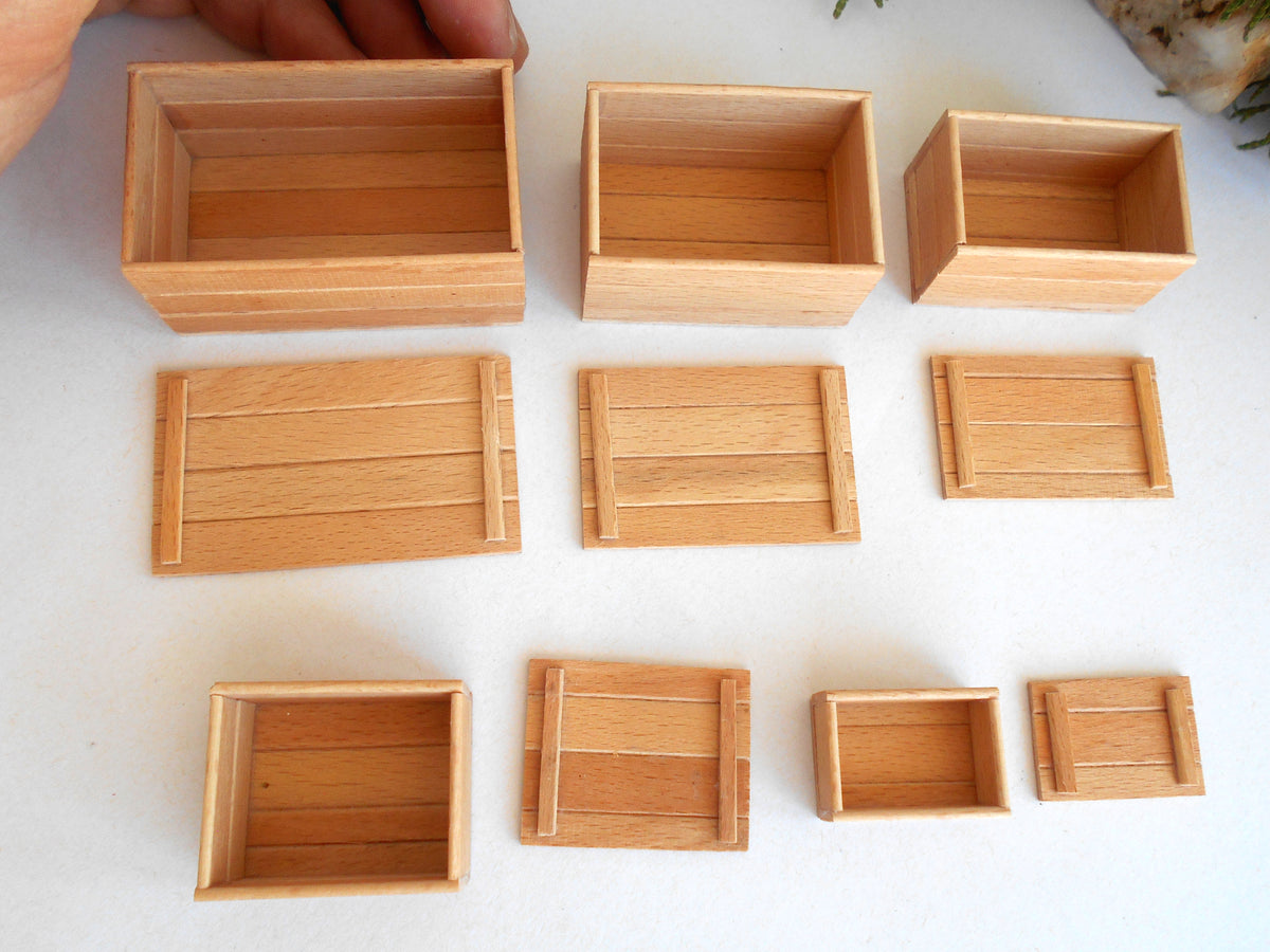 Nine miniature light oak wood boxes of various sizes and shapes, including some with slatted fronts, arranged on a white surface. A hand is visible holding one of the larger boxes at the top left.